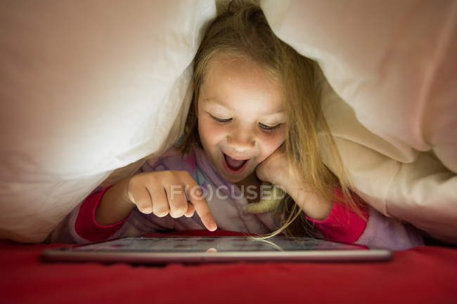 Muchacha preescolar emocionada usando tableta debajo de la manta en la cama - foto de stock