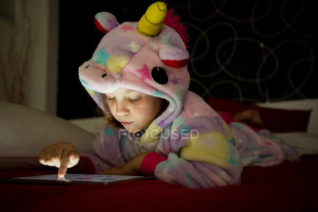 Girl in unicorn pajama using tablet on bed in evening — Stock Photo