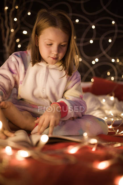 Adorable preschooler girl in soft cozy pajama sitting on bed decorated with Christmas lights and reading book — Stock Photo