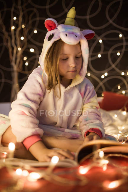 Pequeña chica linda lectura libro en el dormitorio decorado con luces de Navidad - foto de stock