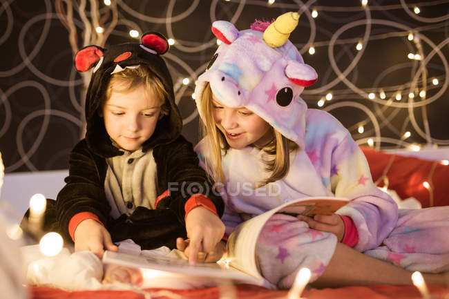 Niedliche Geschwister im kuscheligen Kigurumi-Pyjama lesen Märchenbuch, während sie zusammen auf dem Bett mit Weihnachtsbeleuchtung dekoriert sitzen — Stockfoto