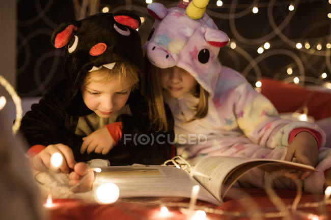Niedliche Geschwister im kuscheligen Kigurumi-Pyjama lesen Märchenbuch, während sie zusammen auf dem Bett mit Weihnachtsbeleuchtung dekoriert sitzen — Stockfoto