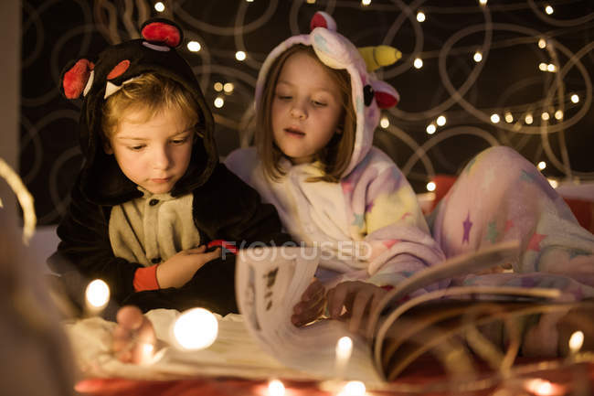 Lindos hermanos en acogedor pijama kigurumi leer cuentos de hadas libro mientras se sientan juntos en la cama decorada con luces de Navidad - foto de stock