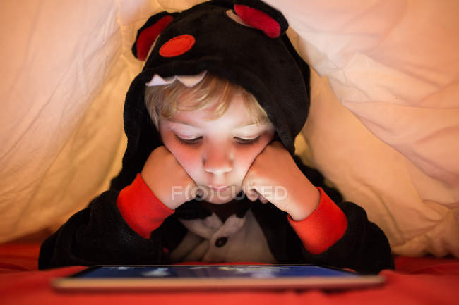 Menino pré-escolar em kigurumi pijama usando tablet sob cobertor na cama — Fotografia de Stock
