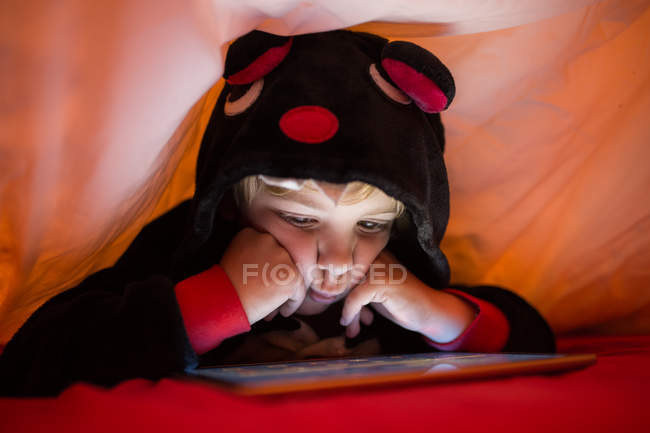 Kleiner Vorschuljunge im kigurumi-Pyjama mit Tablet unter Decke im Bett — Stockfoto