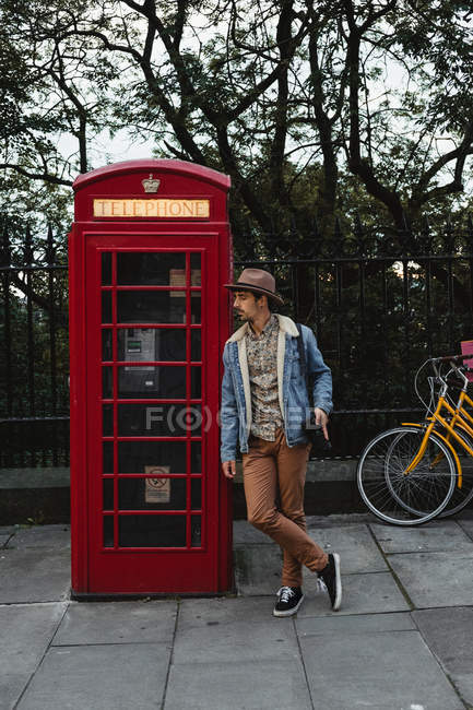 Relaxed male photographer in hat leaning on traditional old style telephone box against green trees on street in Scotland — Stock Photo