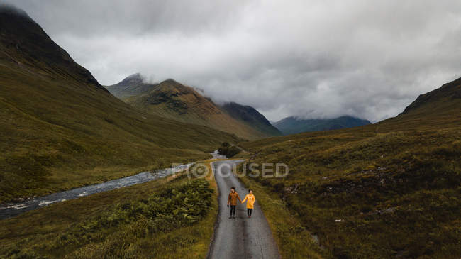 Desde arriba pareja disfrutando de la vista en el camino vacío y tomados de la mano cerca de río de montaña en verdes prados exuberantes en Escocia - foto de stock