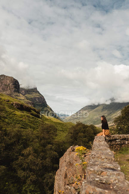 Висококутний вид спокійної жінки, яка сидить на кам'яному паркані і споглядає проти зелених високогір'їв Шотландії. — стокове фото