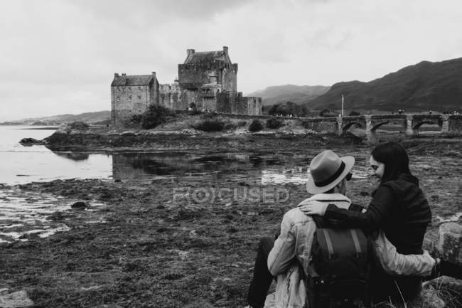 Чорно-біла щаслива пара, яка дивиться один на одного, сидячи на березі проти середньовічного замку в Шотландії. — стокове фото
