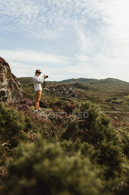 Side view of male traveler with camera taking photo of landscape of Scottish highlands with white clouds in sky — Stock Photo