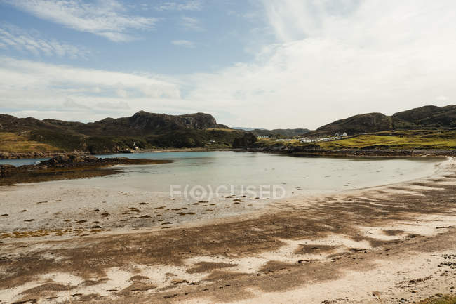 Belo lago com praia de areia cercada por montanhas cênicas e colinas cobertas com grama verde nas terras altas escocesas — Fotografia de Stock