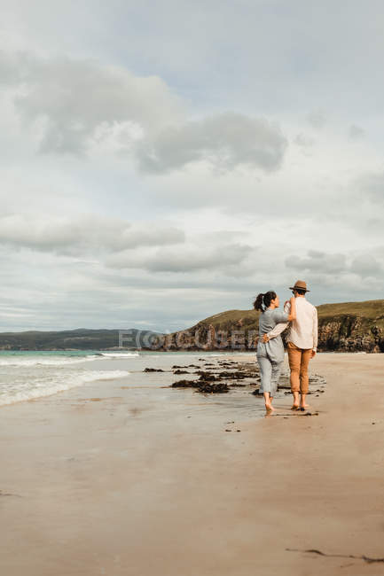 Pareja feliz caminando y abrazando en la costa arenosa escocesa - foto de stock
