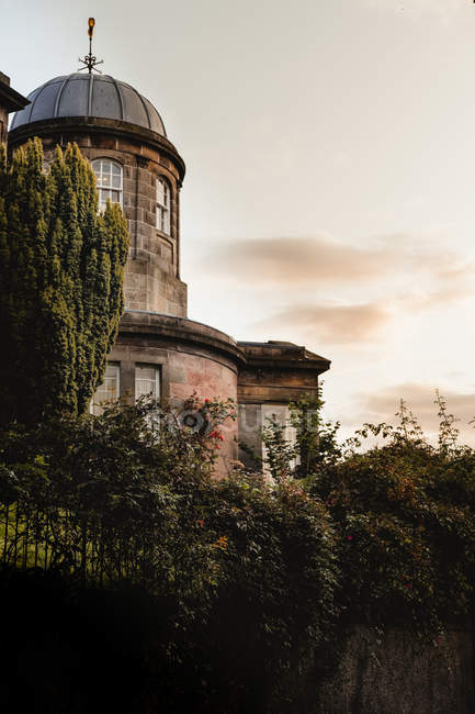 Знизу старої історичної будівлі з куполом в оточенні зелених рослин проти заходу сонця з хмарами в Шотландії. — стокове фото