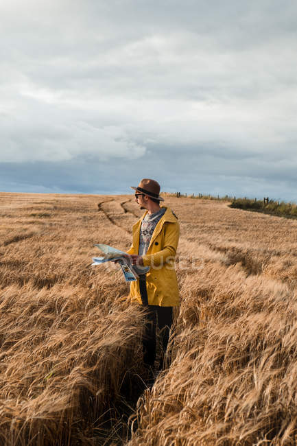 Modern male tourist in yellow jacket and hat standing with map and looking away on footpath in boundless Scottish field — Stock Photo