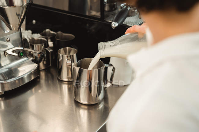 Cropped of female barista pouring milk from glass bottle to metal pot while working at counter with coffee equipment in cafe — Stock Photo