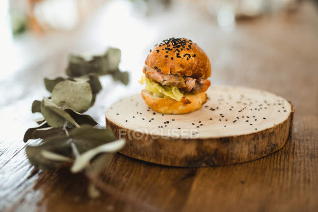Hamburguesa deliciosa con pan crujiente y sésamo negro servido en bandeja redonda de madera sobre mesa de madera con planta - foto de stock