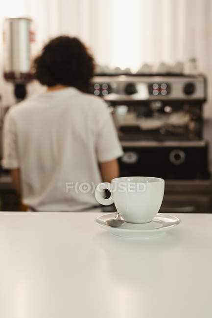 Taza blanca de café en el mostrador de la cafetería con barista femenina borrosa usando la máquina de café en el fondo - foto de stock