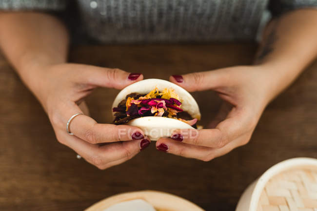 Руки женщины, держащей традиционный азиатский сэндвич с мясом и овощами за столом с бамбуковым пароходом — стоковое фото
