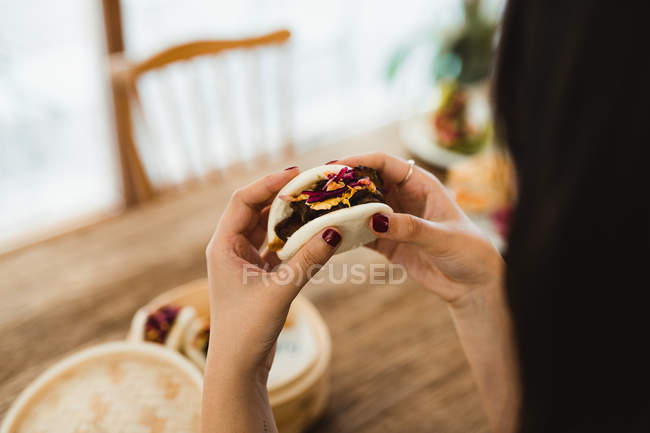 Hands of woman holding traditional Asian steamed sandwich with meat and vegetables over table with bamboo steamer — Stock Photo