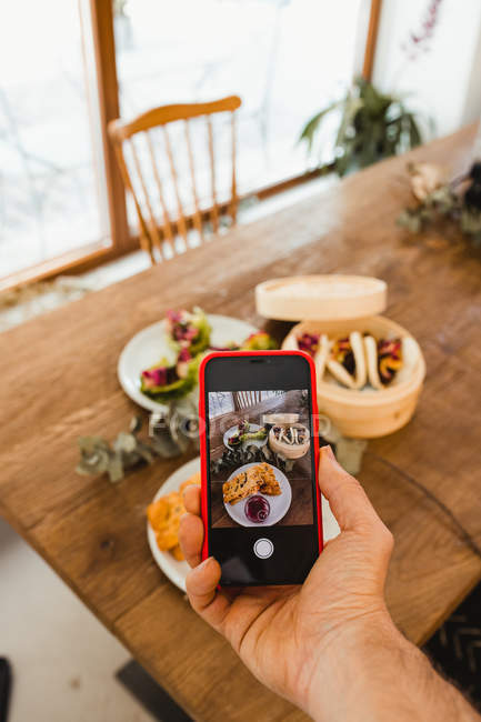 De cima da mão da pessoa que usa o smartphone e tira a foto de vários pratos colocados em conjunto na mesa de madeira — Fotografia de Stock