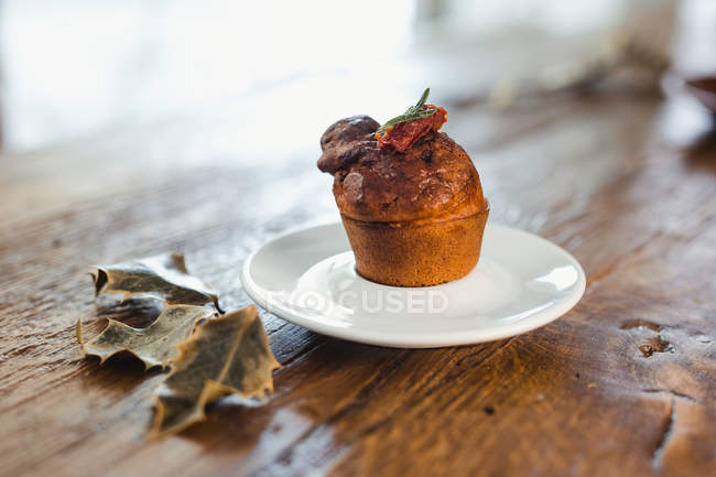 Placa blanca con sabroso muffin recién horneado con hierbas y tomates secos sobre mesa de madera con hojas - foto de stock