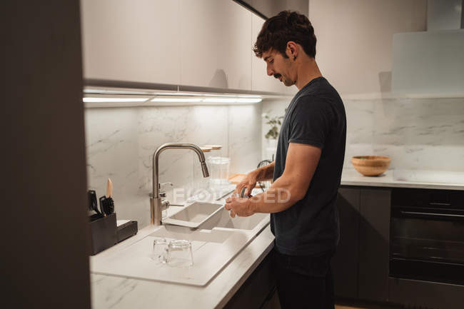 Vue latérale du jeune homme en vêtements décontractés laver la vaisselle dans la cuisine à la maison — Photo de stock