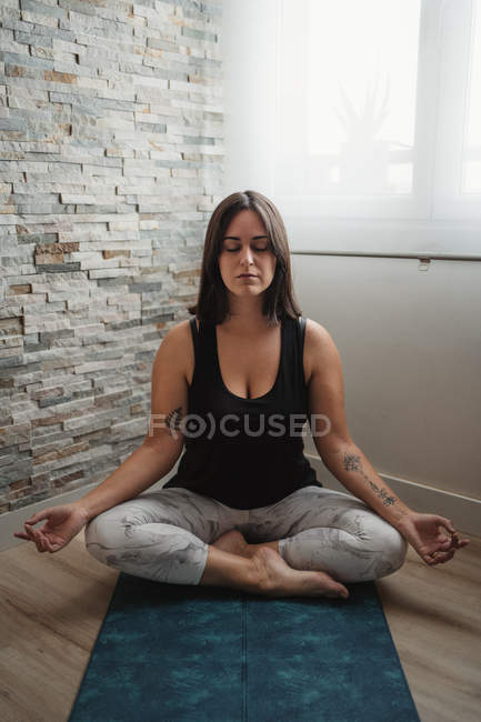 Mujer joven sentada en posición de loto sobre esterilla de yoga y meditando mientras practica yoga matutino en casa - foto de stock