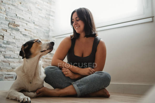 Щаслива молода жінка сидить з схрещеними ногами на підлозі з милим доброзичливим вірним собакою — стокове фото