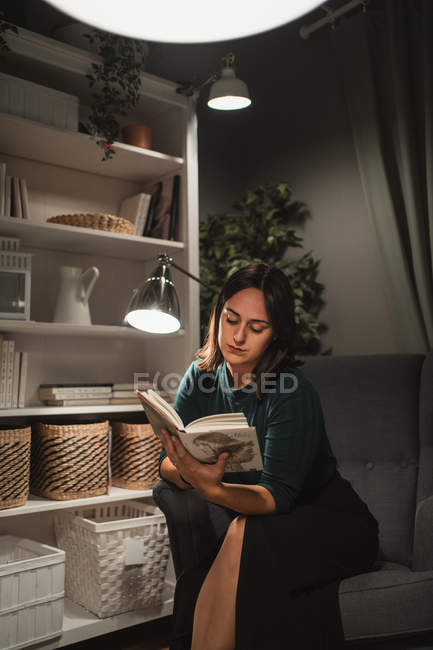 Junge elegante Frau sitzt im Stuhl im gemütlichen Zimmer neben Lampe und liest Buch mit Lieblingsroman, während zu Hause ausruhen — Stockfoto