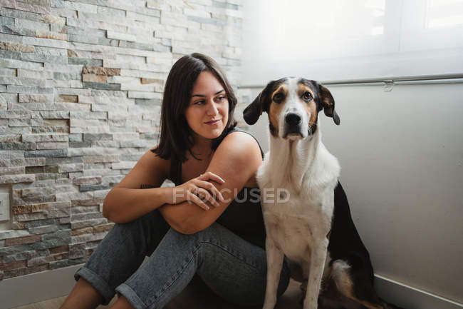 Молода весела жінка сидить з доброзичливою змішаною собакою на підлозі вдома — стокове фото