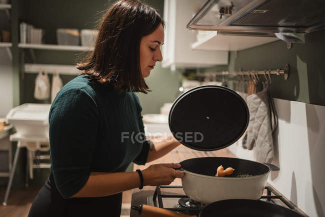 Vista lateral da jovem fêmea removendo a tampa da panela e verificando a comida enquanto estava no fogão e preparando o jantar em casa — Fotografia de Stock