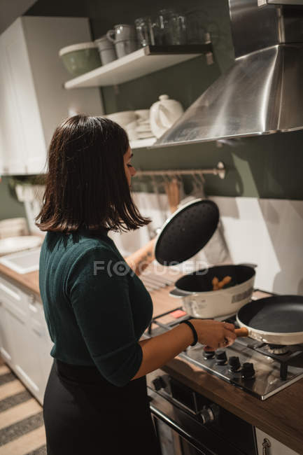 Вид спереду молодої жінки, що знімає кришку сковороди та перевіряє їжу, стоячи на плиті та готуючи вечерю вдома — стокове фото