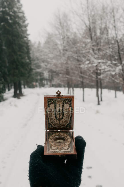 Mano de recorte de viajero en guante de punto negro con brújula vintage con reloj de sol mientras está de pie en el camino nevado entre el bosque en el sombrío día de invierno - foto de stock