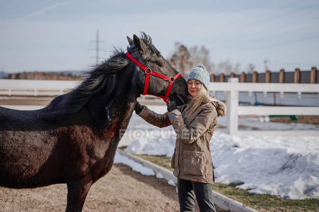 Вид сбоку лошади в заснеженном дворе и женщины в теплой шляпе и куртке в яркий холодный день — стоковое фото