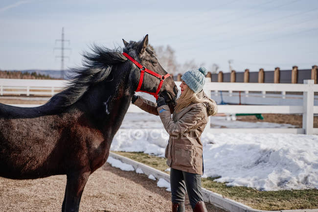 Side view of luxurious horse in snowy yard and woman in warm hat and jacket in bright cold daylight — Stock Photo