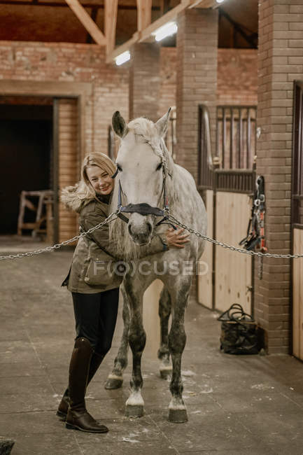 Довгошерста блондинка обіймає сірого коня з білою мордою гри і дивиться в камеру в стайні — стокове фото