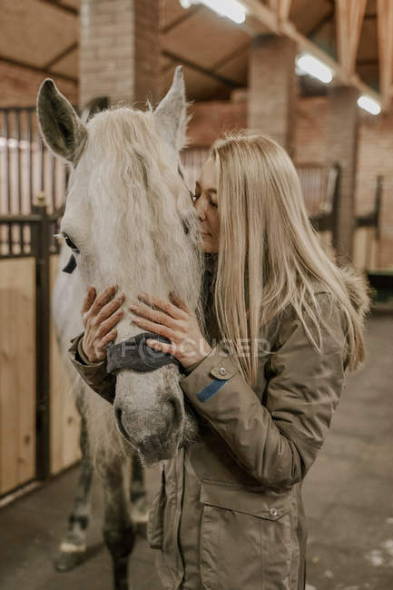 Mujer de pelo largo abrazando caballo gris manzana con bozal de melena blanca en establo - foto de stock