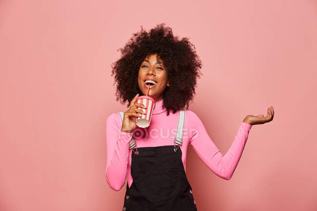 Heureuse femme noire avec une tasse de boisson jetable debout sur fond rose — Photo de stock