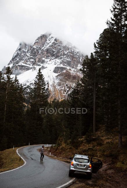 Lonely road with car on roadside and traveler among pine forest near big cliffs — Stock Photo