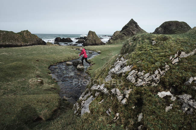 De cima mulher anônima desfrutando paisagem cênica incrível da Irlanda do Norte durante a viagem enquanto caminhava perto do rio raso rápido que flui para águas de costa rochosa — Fotografia de Stock