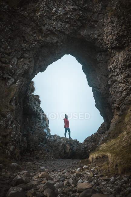 Vista lateral de la hembra en traje caliente de pie en el borde del acantilado dentro de la cueva en el puerto de Irlanda del Norte mirando hacia el mar - foto de stock