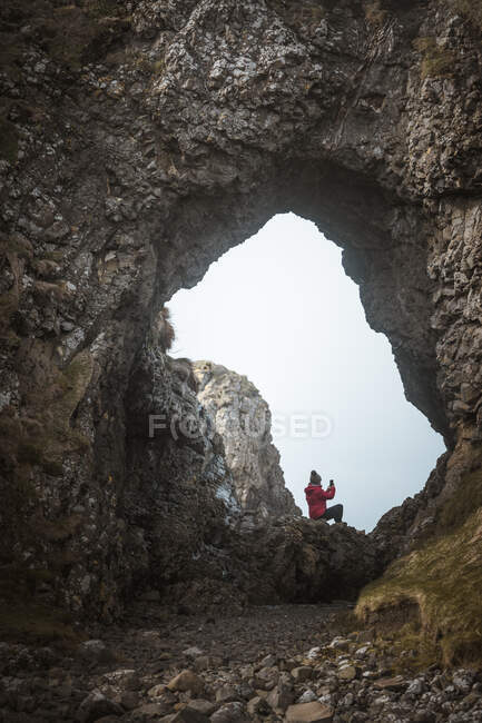 Vue arrière de la femelle en tenue chaude assise sur le bord de la falaise dans la grotte dans le port de l'Irlande du Nord regardant loin dans la mer en utilisant un téléphone mobile — Photo de stock