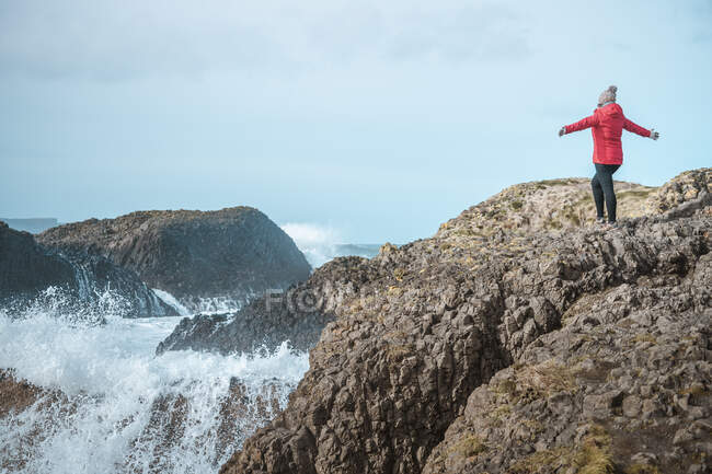 Back view of faceless free female in warm outfit on cliff top enjoying stormy raging surf with spread arms in Ballintoy — Stock Photo