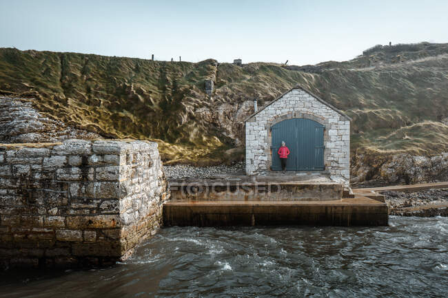 Feminino turista em roupa quente apoiando-se no antigo edifício de pedra com portas, enquanto em pé no pitoresco cais de Ballintoy porto sobre rochas fundo — Fotografia de Stock