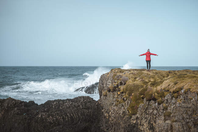 Back view of faceless free female in warm outfit on cliff top enjoying stormy raging surf with spread arms in Ballintoy — Stock Photo