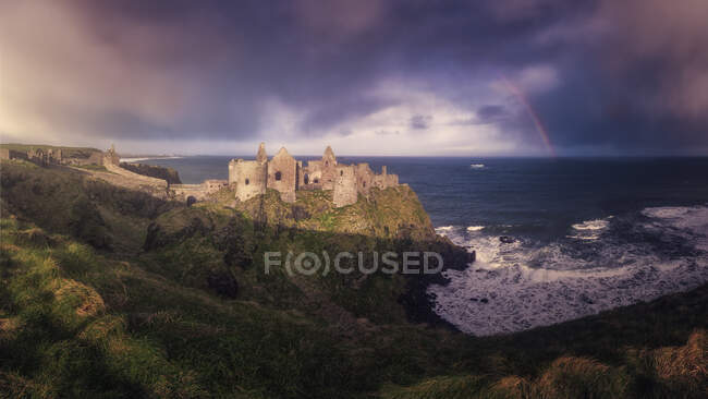 Majestic landscape with ruins of ancient stone Dunluce Castle in sunlight located on edge of cliff with rainbow breaking through stormy sky and sea waves beating against shore — Stock Photo