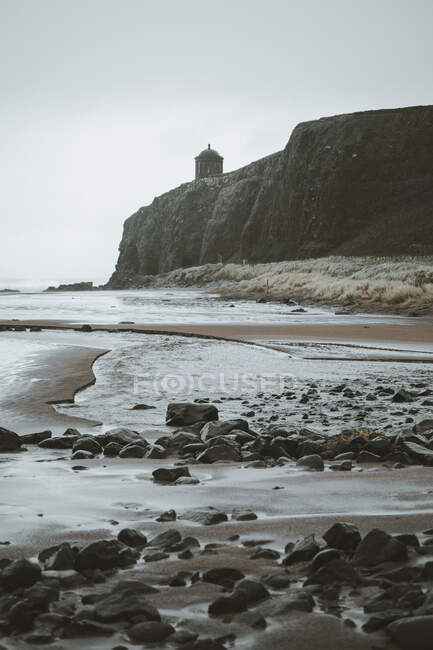 Paisaje escénico con el templo de Mussenden situado en el acantilado de piedra en la costa de Irlanda del Norte y olas de mar tormentosas que se estrellan contra las rocas con el cielo gris nublado en el fondo - foto de stock