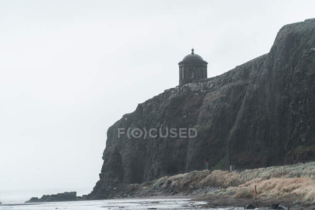 Сценический пейзаж с храмом Муссендена, расположенным на каменной скале у береговой линии Северной Ирландии, и бурные морские волны, ударяющиеся о скалы с серым облачным небом на заднем плане — стоковое фото