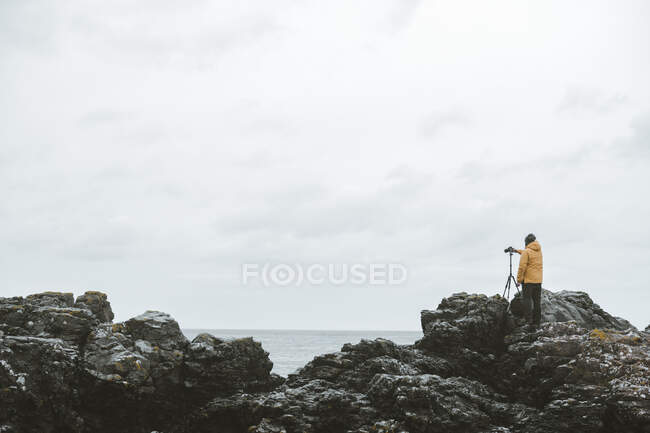 Vista posteriore del viaggiatore di sesso maschile in piedi sulla roccia con fotocamera su treppiede e scattare foto del paesaggio marino in giornata nuvolosa cupa sulla costa dell'Irlanda del Nord — Foto stock