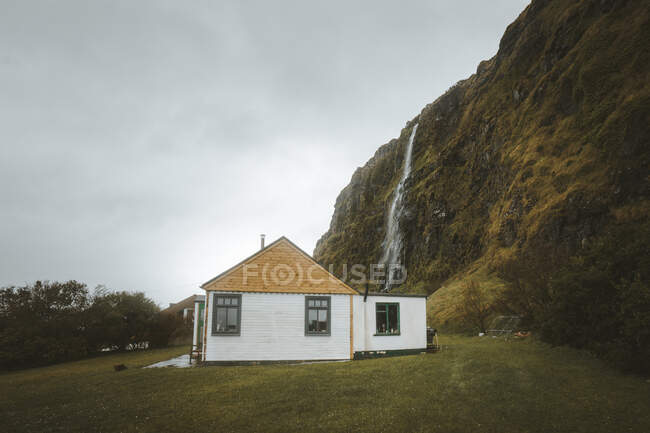 Маленький деревенский деревянный дом с белыми стенами и желтой крышей, расположенный на зеленом подножии скалы с водопадом на фоне серого облачного неба в весенний день в Северной Ирландии — стоковое фото